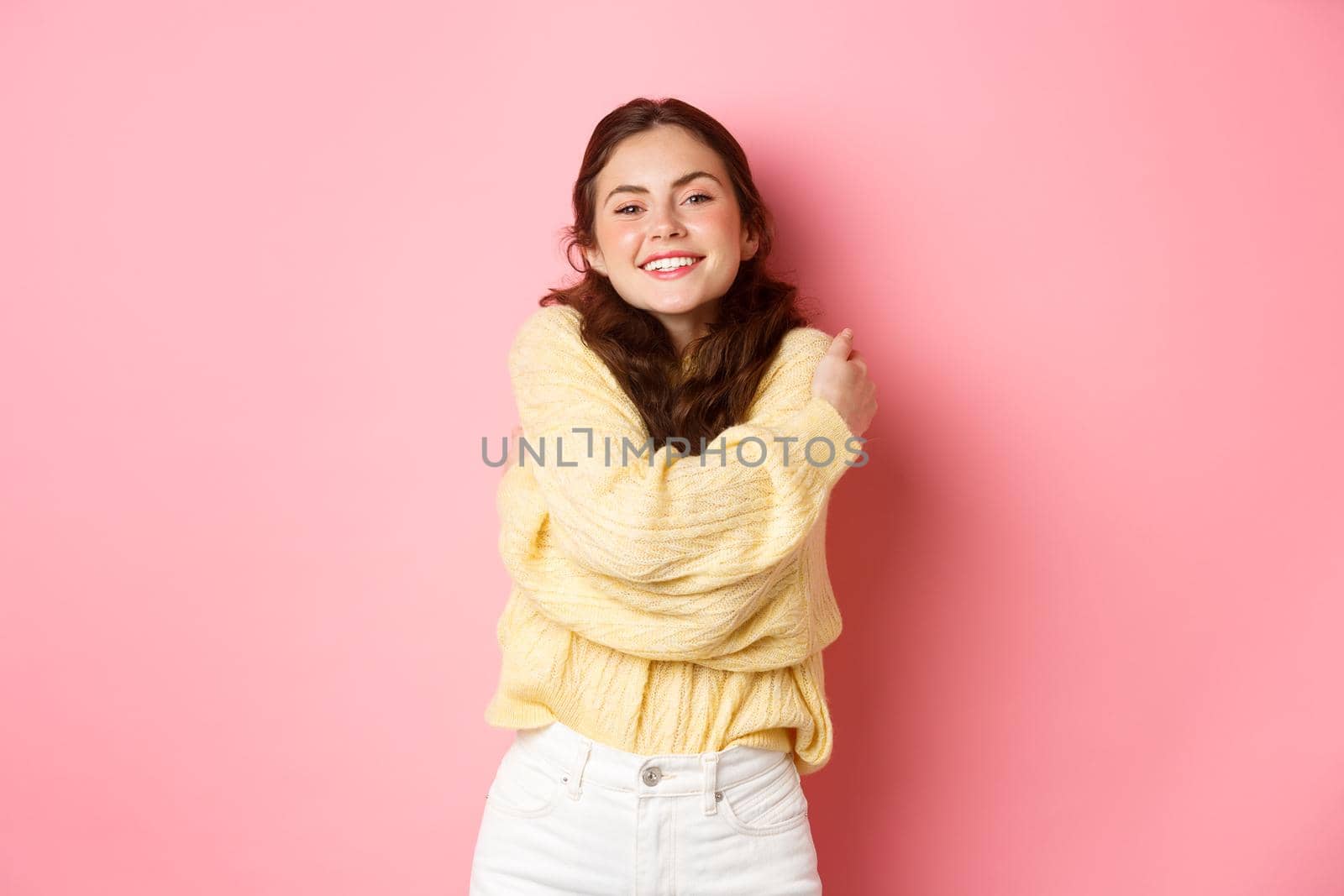 Young beautiful girl feeling healthy and happy, hugging herself while wearing soft sweater, smiling pleased, standing romantic against pink background.