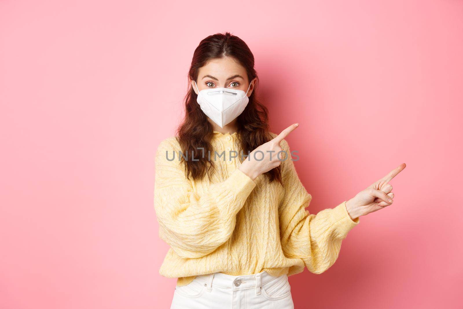 Covid, corona and social distancing concept. Excited female model in respirator shows you advertisement, points at logo aside, wears medical respirator on face, pink background.