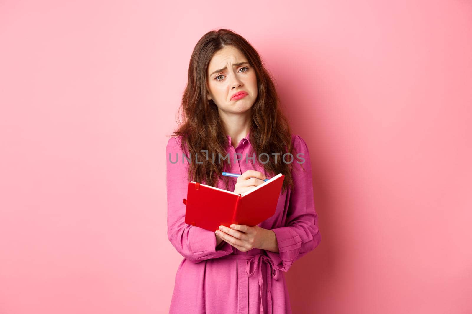 Sad young woman writing her feelings in diary, looking upset and gloomy, feeling heartbroken, sharing memories in planner, standing against pink background.