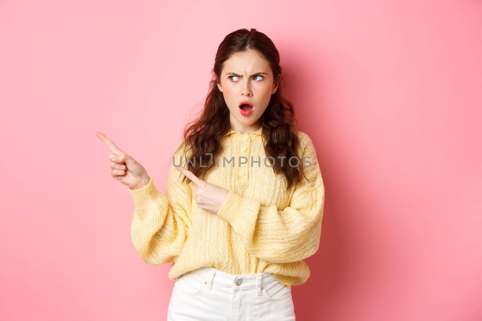 Offended and shocked young woman staring and pointing fingers left at copyspace aside, insulted by something, gasping displeased, standing against pink background.