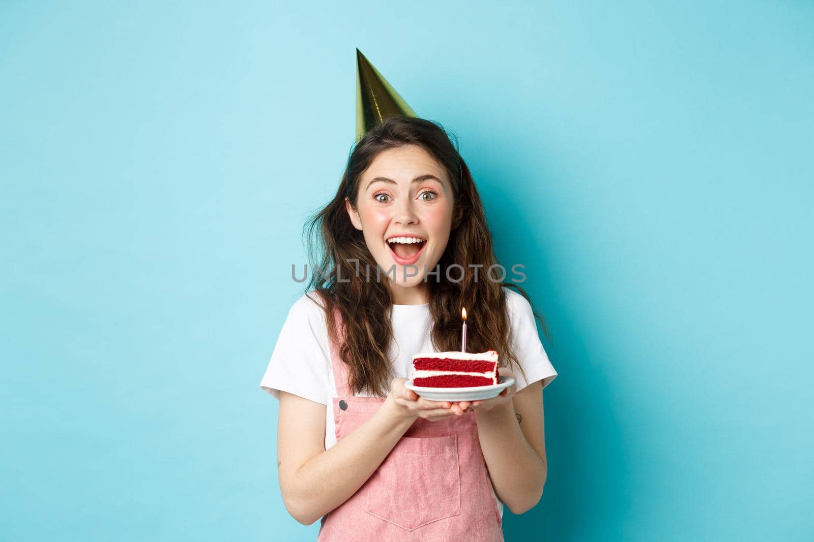 Holidays and celebration. Cheerful birthday girl in party hat holding bday cake and smiling, making wish on lit candle, standing against blue background.