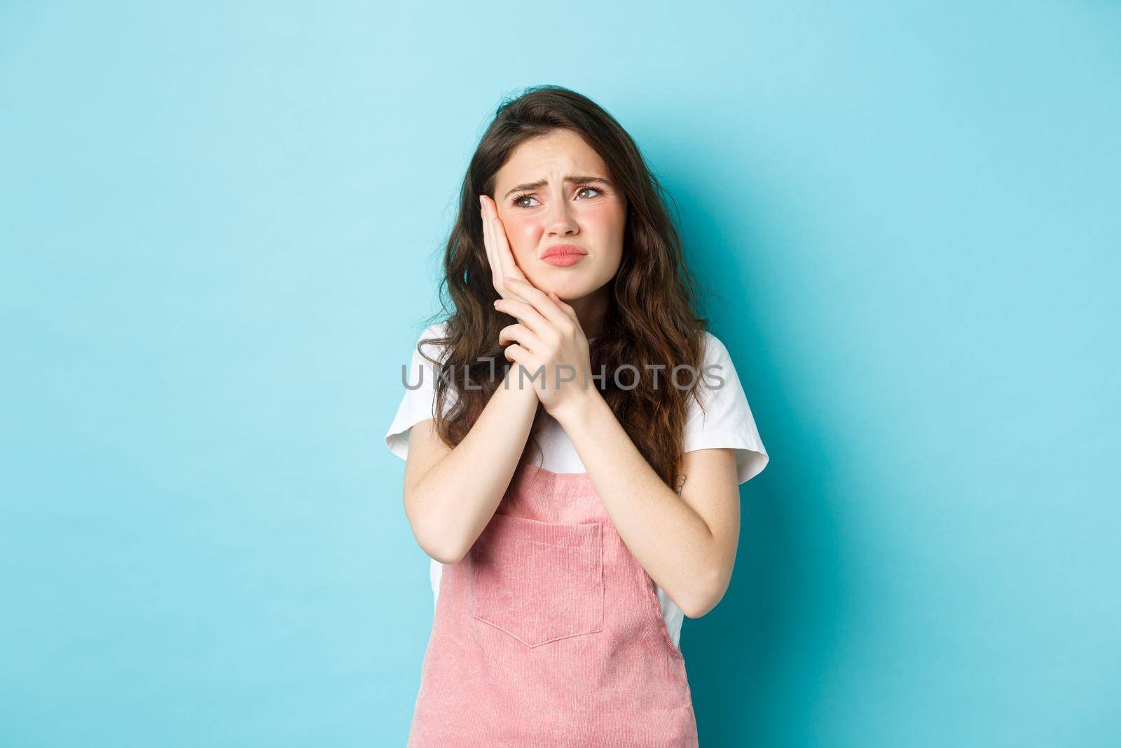 Portrait of frowning girl touching cheek, having toothache, looking at upper left corner stomatology clinic banner, suffering tooth decay, standing against blue background.