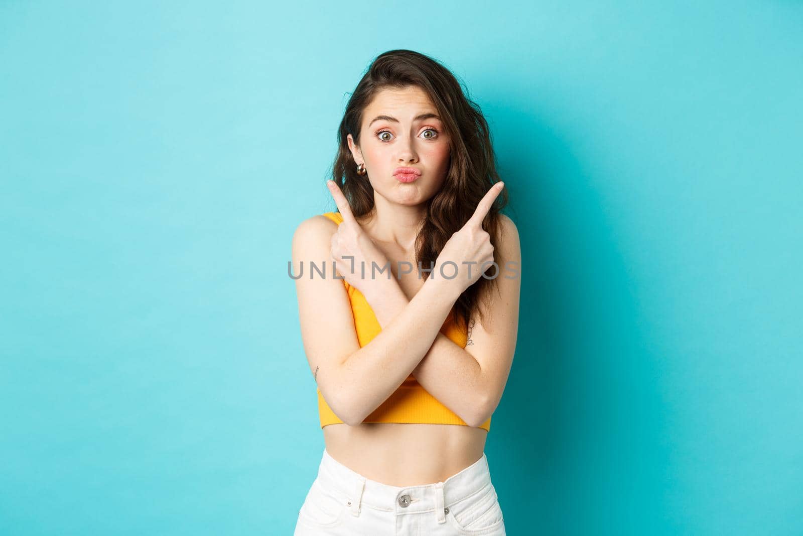 Young confused woman pointing sideways, need help with choice, showing two variants and look indecisive, standing over blue background.
