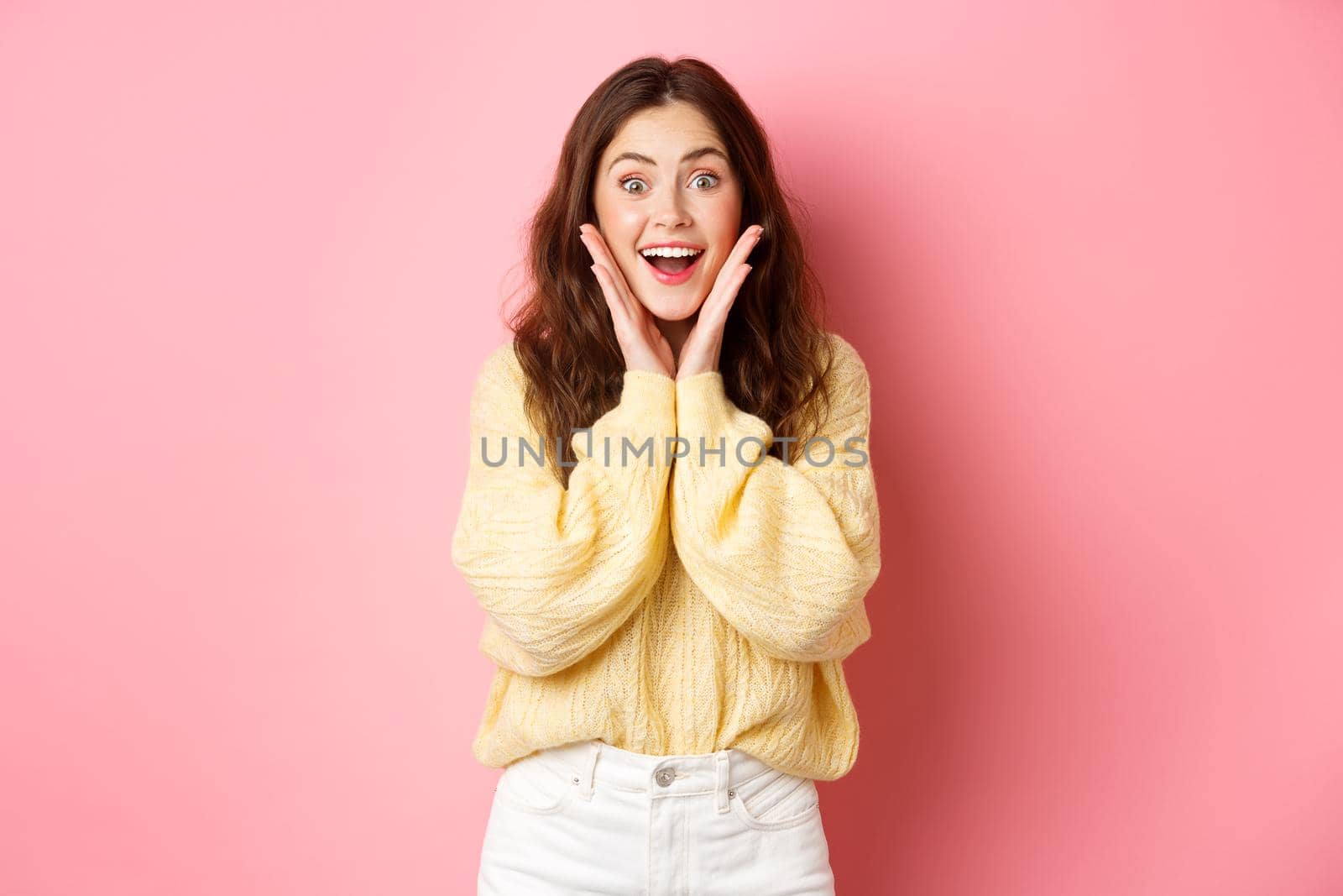 Excited beautiful girl staring surprised and amazed, holding hands on cheeks and gasping, looking with astonished face at promotional offer, standing near copy space pink background.