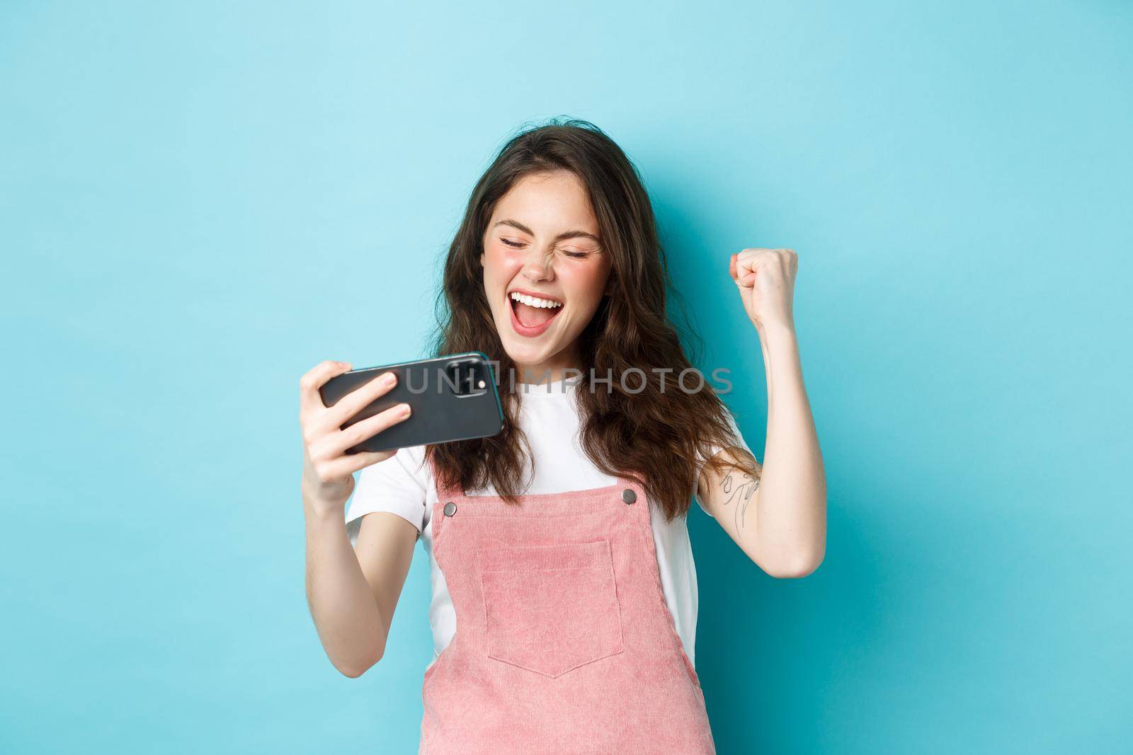 Cheerful cute girl winning in online video game on smartphone, making fist pump and shouting yes with joy, standing over blue background and triumphing by Benzoix