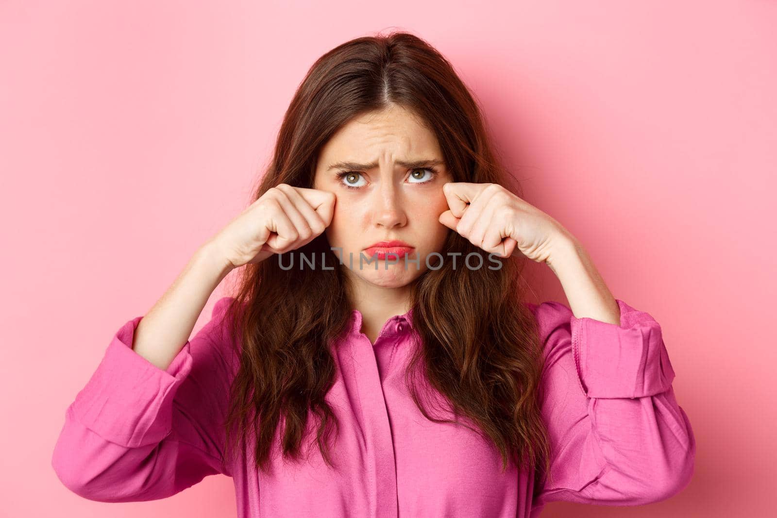 Close up portrait of gloomy young woman crying, wiping tears and sulking from something unfair, frowning and looking up sad, standing over pink background.