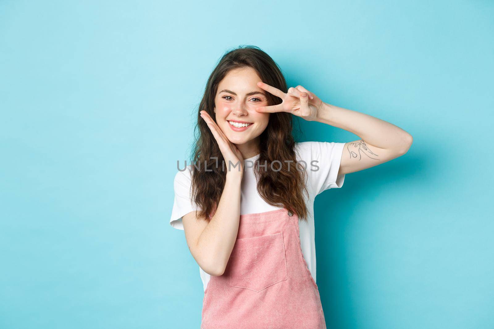 Portrait of kawaii teenage girl giggle and showing peace v-sign near eye, smiling cheerful and touching cheek, standing over blue background.
