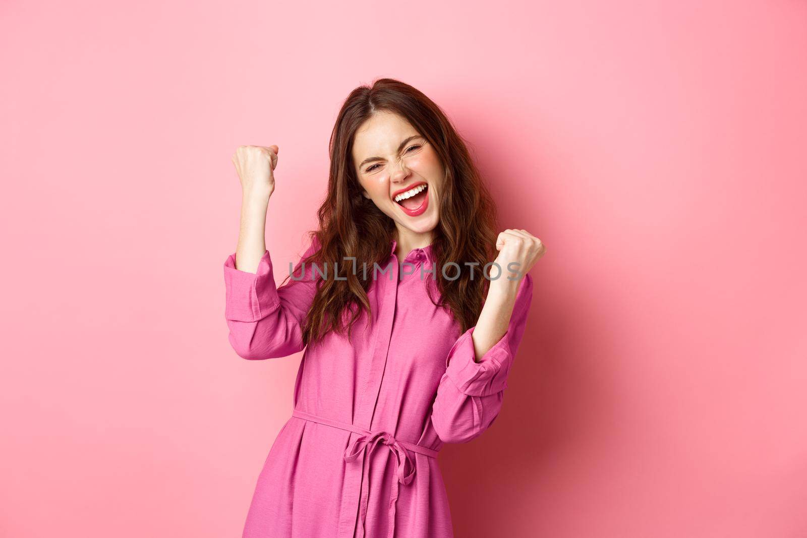 Beautiful smiling woman scream with happy and excited face, saying yes, making fist pump, winning and feeling like champion, triumphing, standing over pink background.