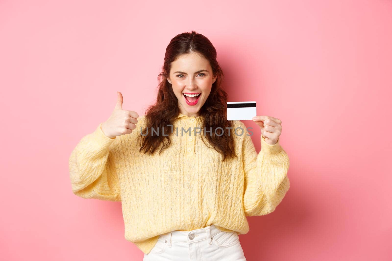Young modern woman saying yes and showing thumbs up, holding plastic credit card, standing over pink background. Copy space