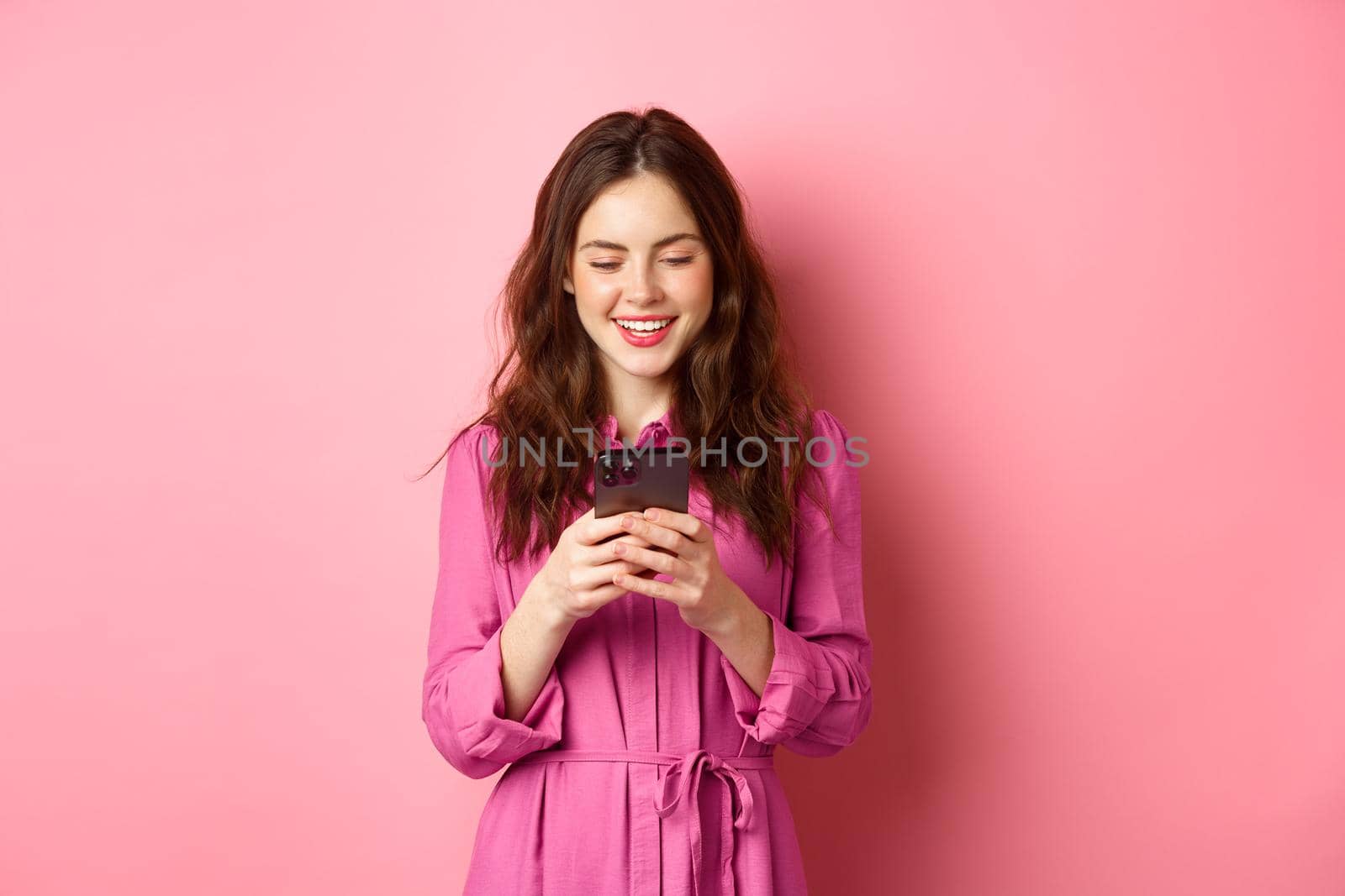 Young woman shopping, chatting on mobile phone, smiling while reading smartphone screen, standing against pink background. Copy space