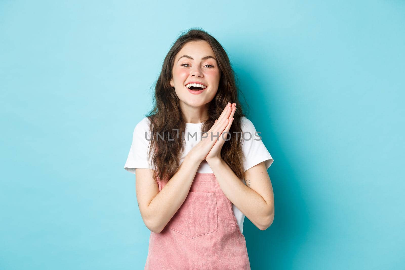 Portrait of happy woman rejoicing of good news, clap hands from dealight and smiling, thanking for gift, looking satisfied, standing against blue background.