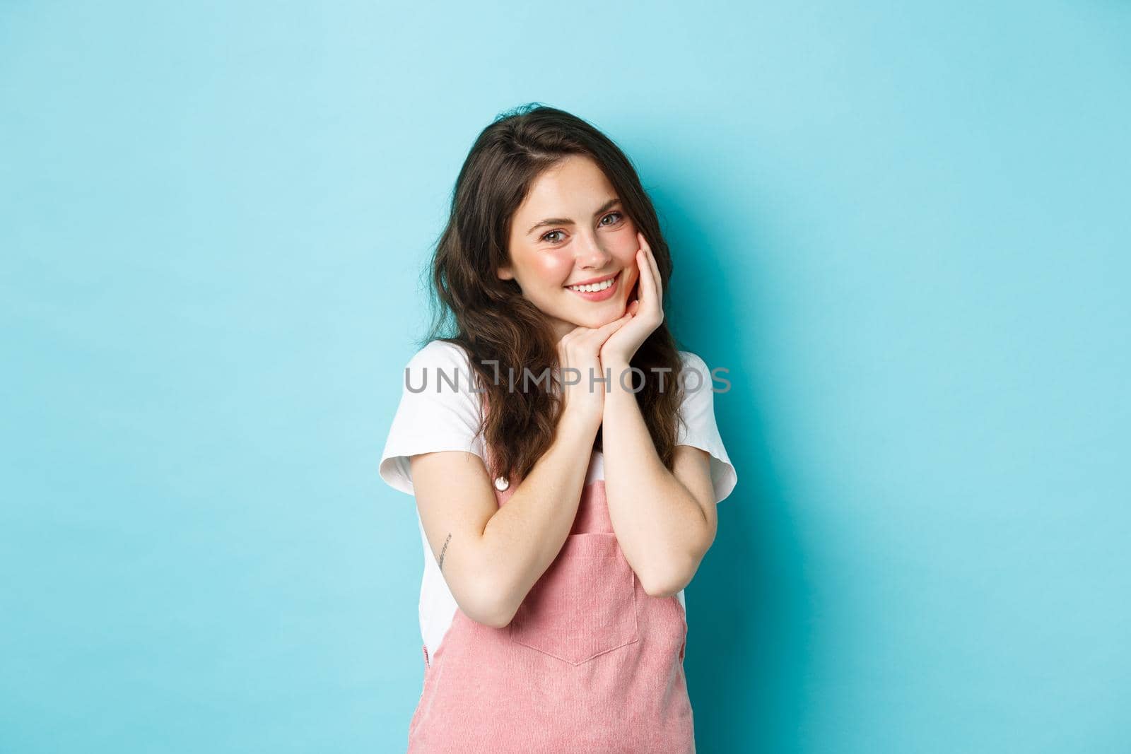 Portrait of silly and cute young woman giggle flirty, smiling and looking coquettish at camera, blushing, having summer outfit and bright make up, standing over blue background.