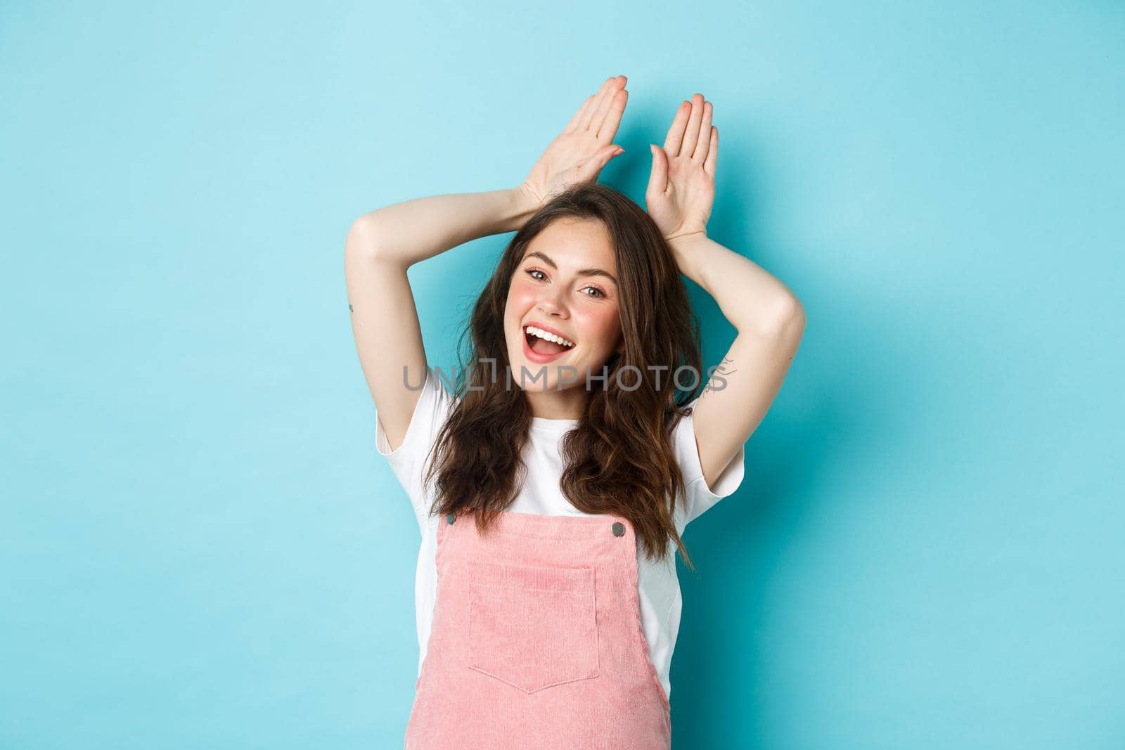Beautiful girl mimic Easter bunny, showing rabbit ears with hands on head and smiling lovely, dancing happy, standing over blue background.