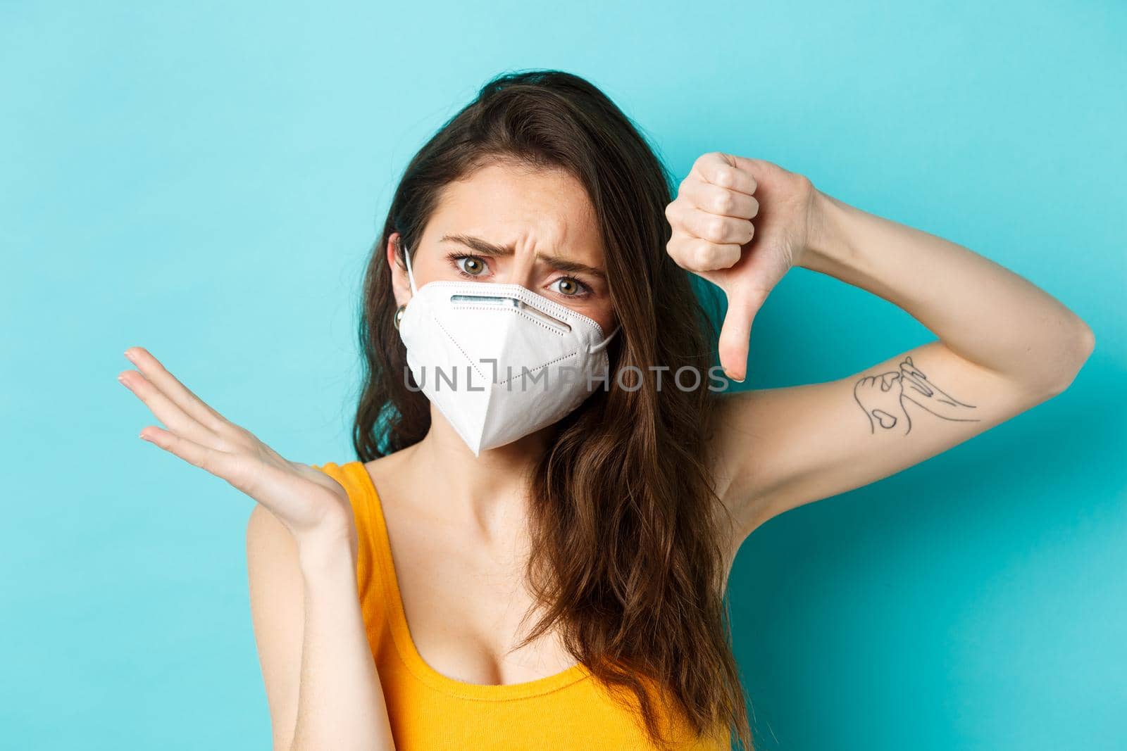 Corona, pandemic and healthcare concept. Frustrated young woman showing thumb down and look confused, dont understand why, wearing face mask, blue background.