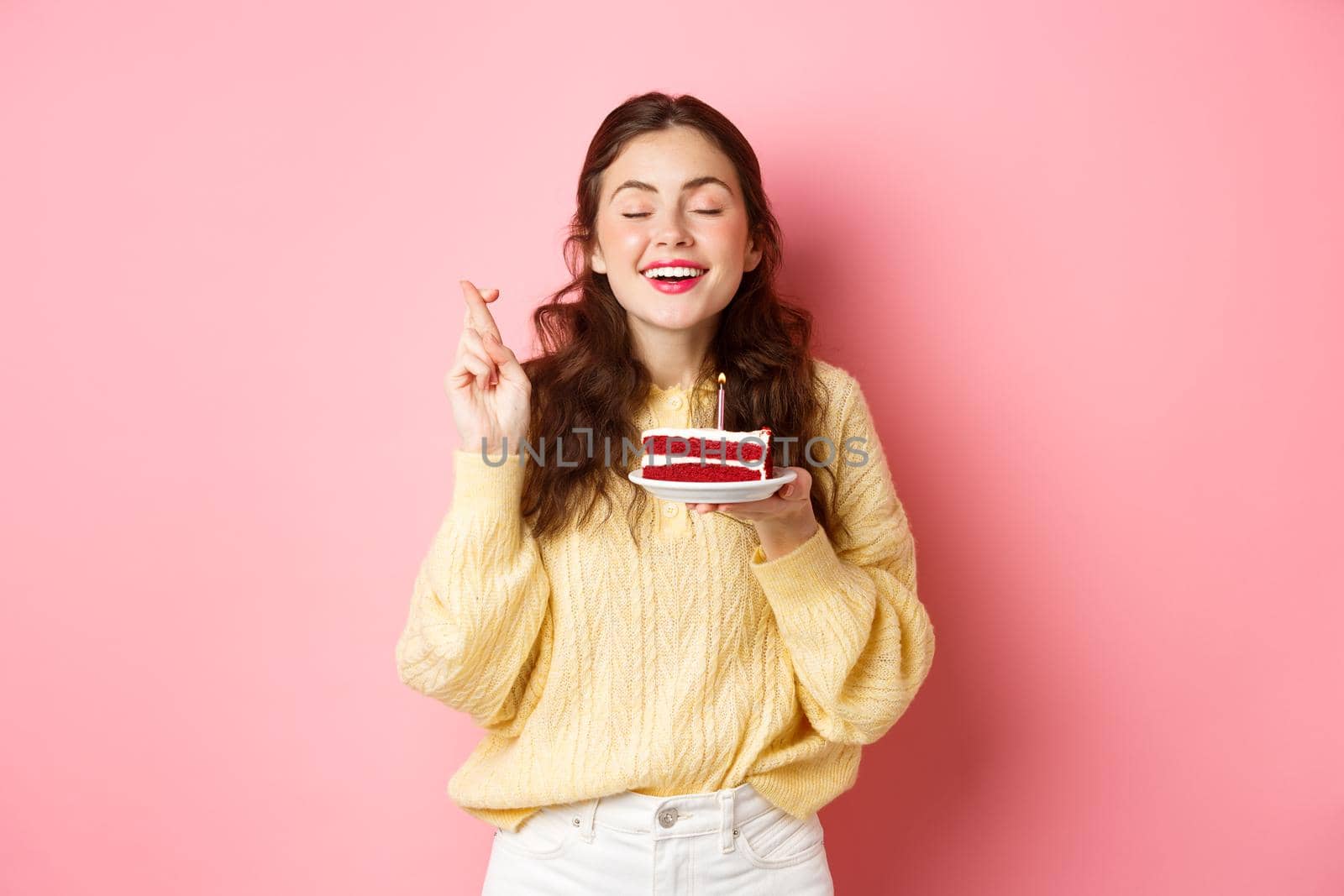 Celebration and holidays. Happy woman celebrates her birthday, makes wish with closed eyes and crossed fingers, holds bday cake with one candle, stands on pink background.