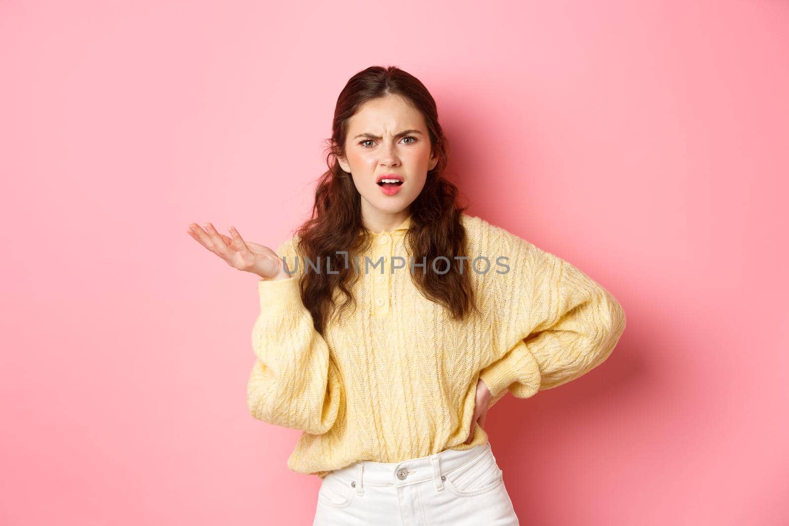 What is this, are you kidding me. Annoyed young woman complaining at something strange, staring confused, raising one hand with puzzled face, standing over pink background.
