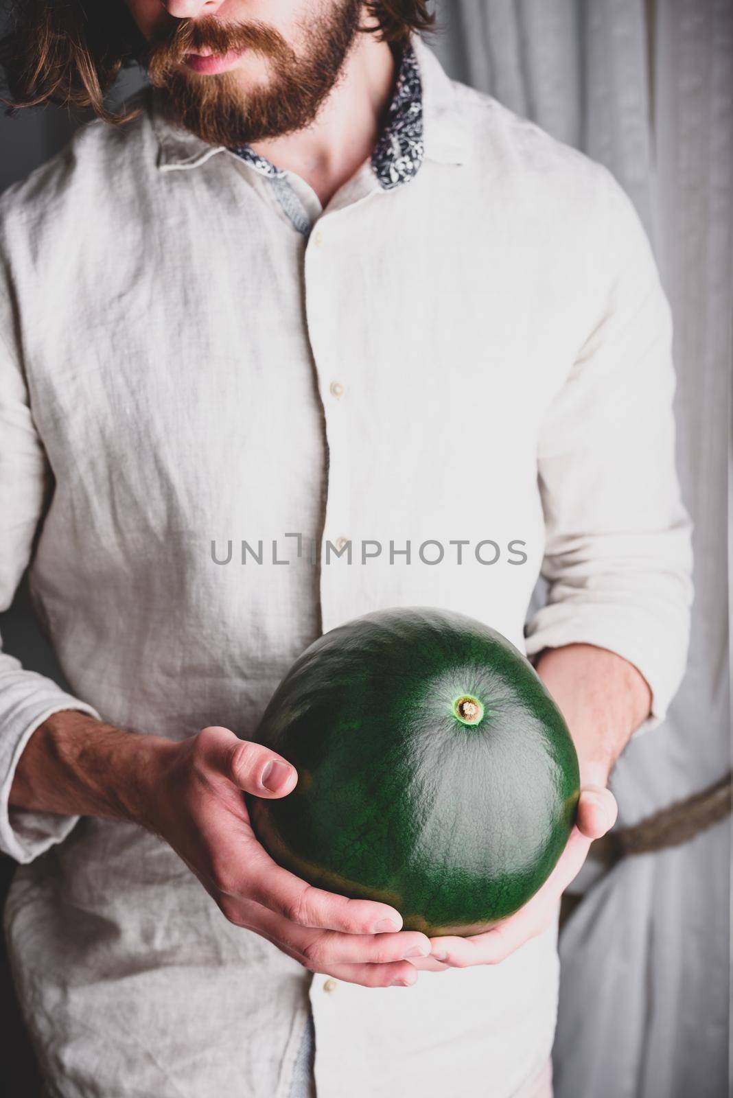 Bearded man in linen shirt holds in his hands whole watermelon