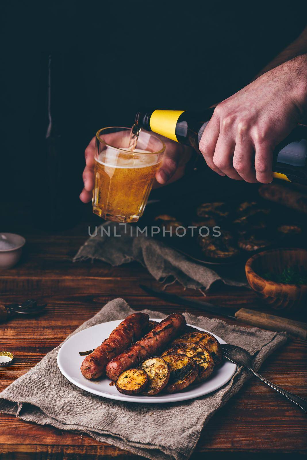 Pork Sausages Baked with Eggplant, Leek and Herbs on White Plate. Man Pouring Beer from Bottle into a Glass.