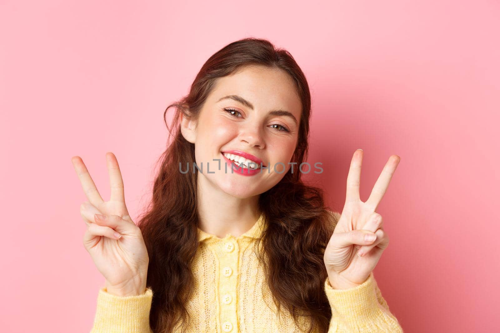 Close up of beautiful young woman showing peace v-sign and smiling happy at camera, wearing bright glam make up, standing against pink background.