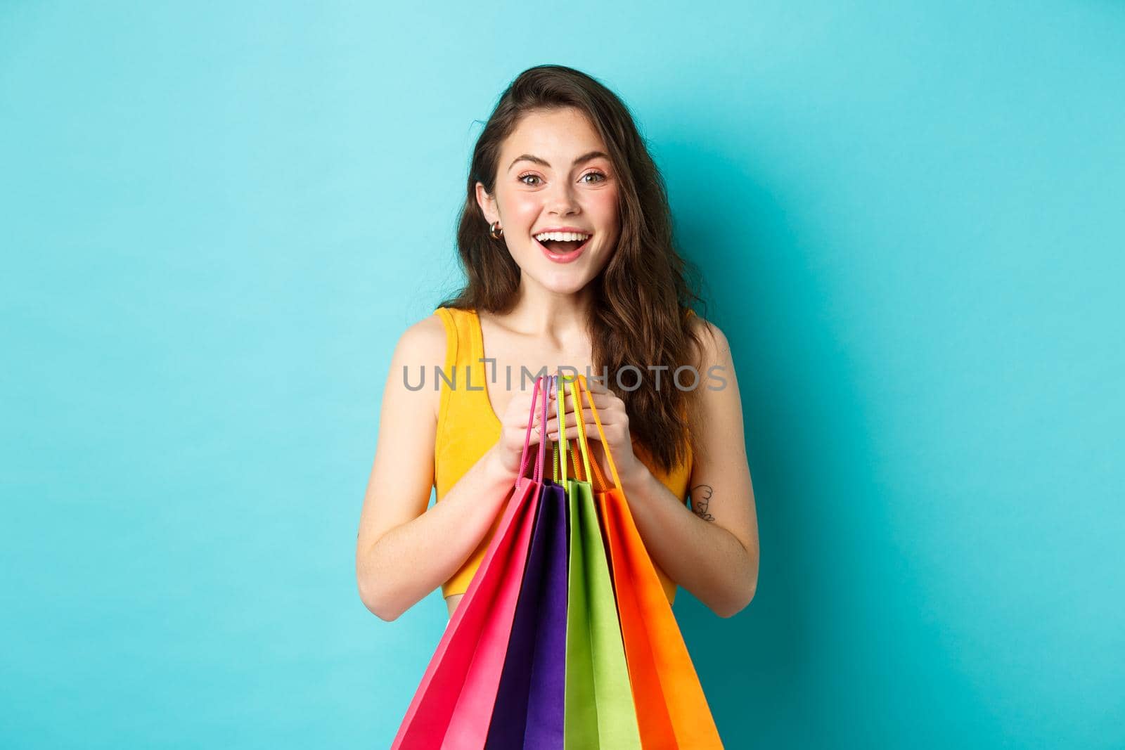 Young pretty woman holding shopping bags, smiling excited at camera, buying with discounts, standing over blue background.