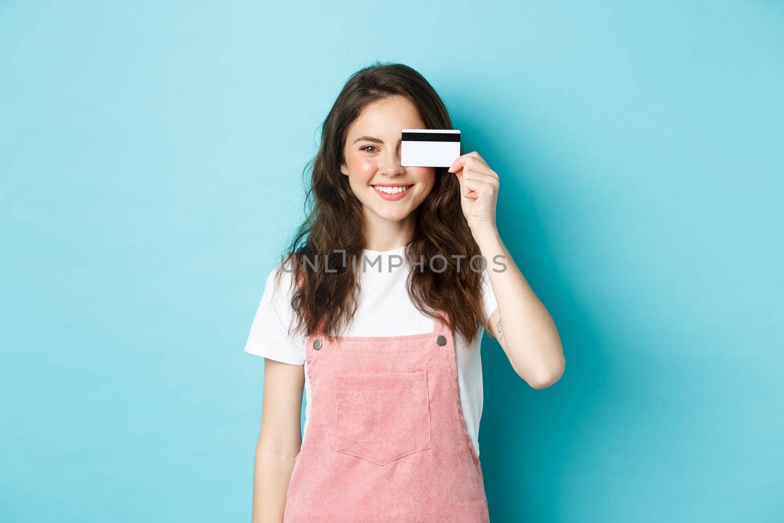 Cute young woman showing plastic credit card on eye and smiling, shopping with it, standing over blue background.