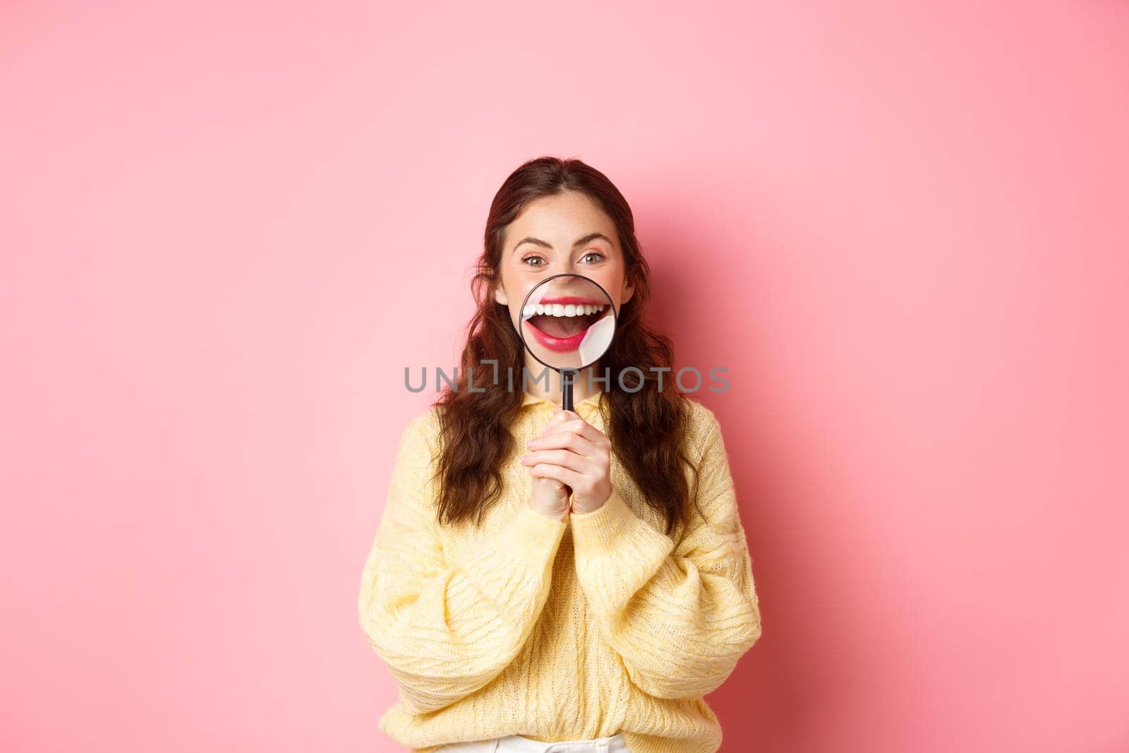 Young smiling woman showing her perfect whitened teeth smile with magnifying glass, promo of dental clinic, stomatology and healthcare concept, standing against pink background.