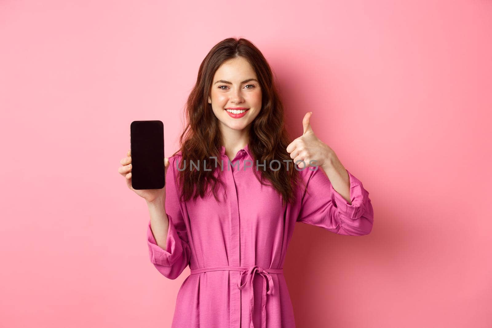 Technology concept. Young girl in dress, shows her smartphone screen and thumbs up, nod in approval, praise good app, standing over pink background.