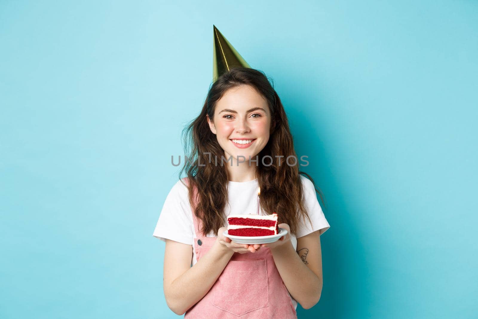 Holidays and celebration. Cheerful birthday girl in party hat holding bday cake and smiling, making wish on lit candle, standing against blue background.