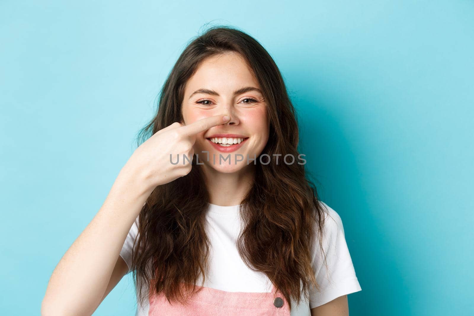 Close up of funny brunette girl touching her nose and smiling cheerfully at camera, standing against blue background.