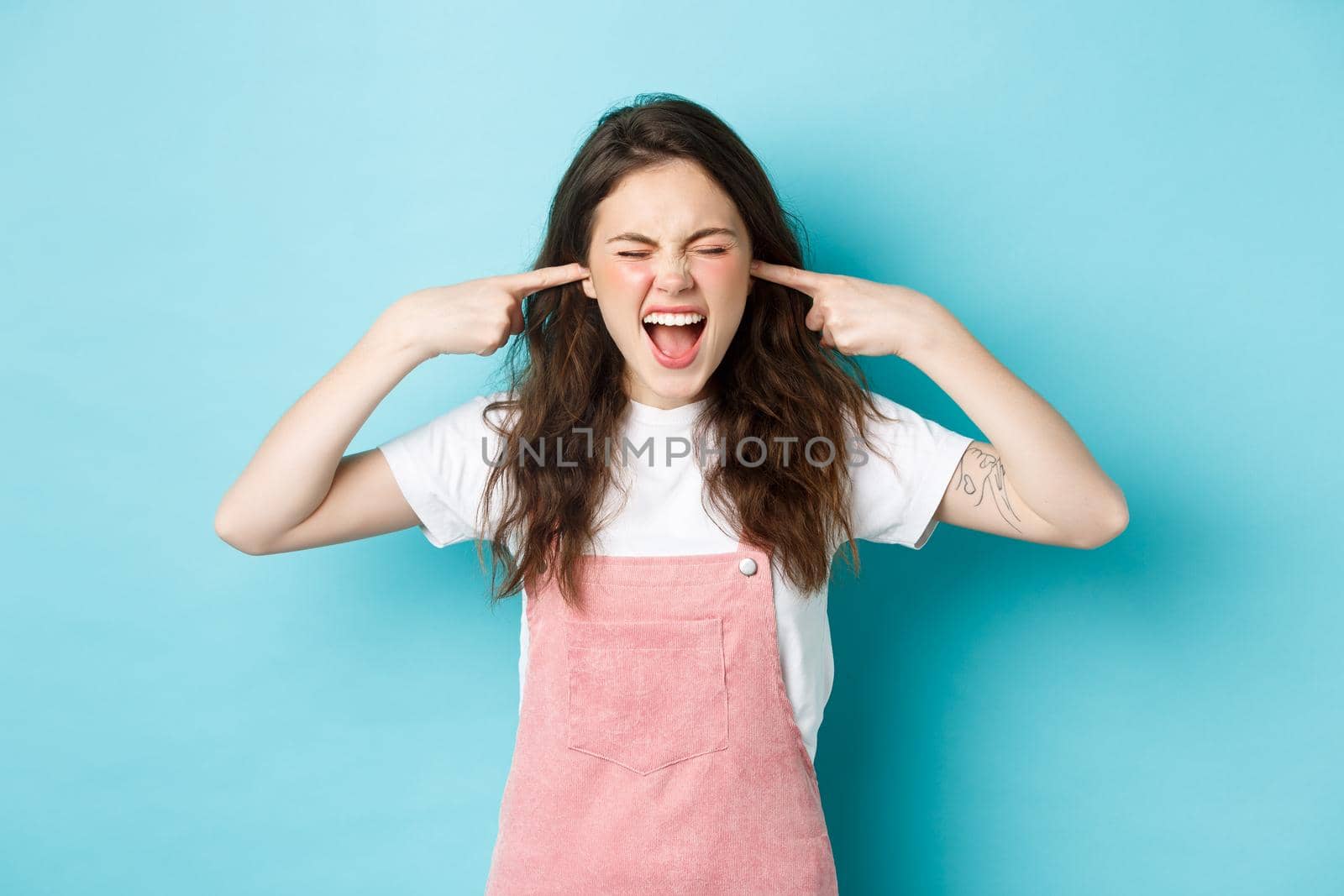 Annoyed young brunette woman screaming and shutting ears, blocking annoying sound, ignoring someone, hear awful noise, standing against blue background.