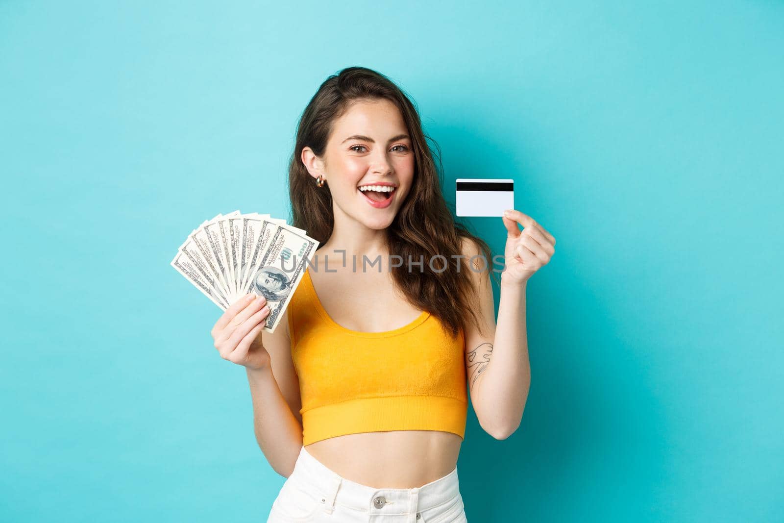 Image of lucky good-looking girl showing plastic credit card and money, holding dollar bills, standing over blue background. Copy space