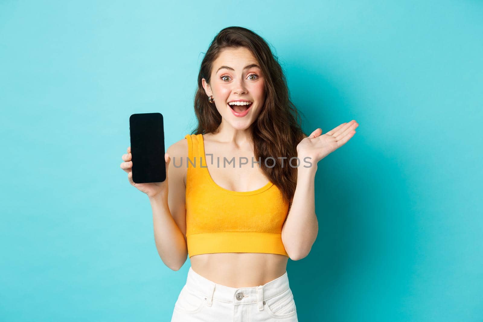 Attractive young woman in glamour summer clothes, showing empty smatphone screen, gasping amazed, recommending app, standing against blue background by Benzoix