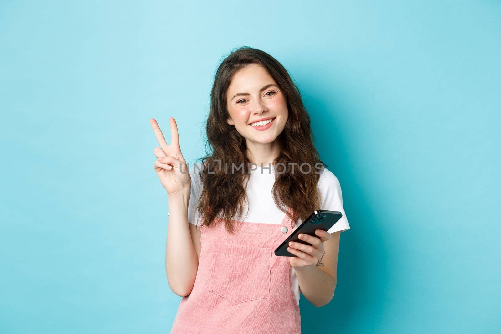 Online shopping. Cute and cheerful young woman smiling, holding smartphone and showing peace v-sign at camera, standing against blue background.