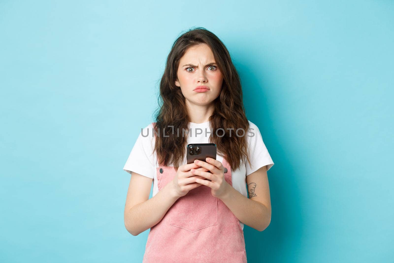 Disappointed girl frowning and grimacing displeased after using smartphone app, holding phone and express dislike, standing over blue background by Benzoix