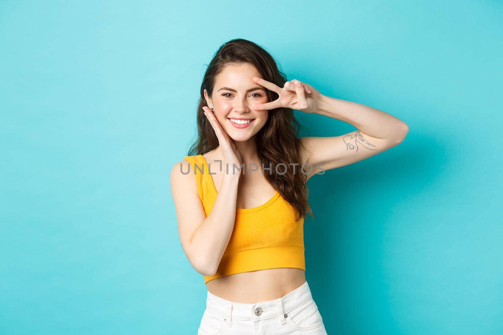 Portrait of beautiful woman with clean skin and perfect body, wearing summer clothes, showing peace sign and touching face, standing over blue background.