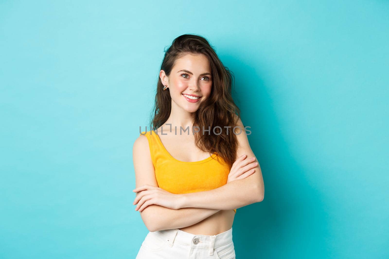 Summer and lifestyle concept. Stylish modern woman in cropped top looking confident, cross arms on chest and smiling delighted, standing over blue background.
