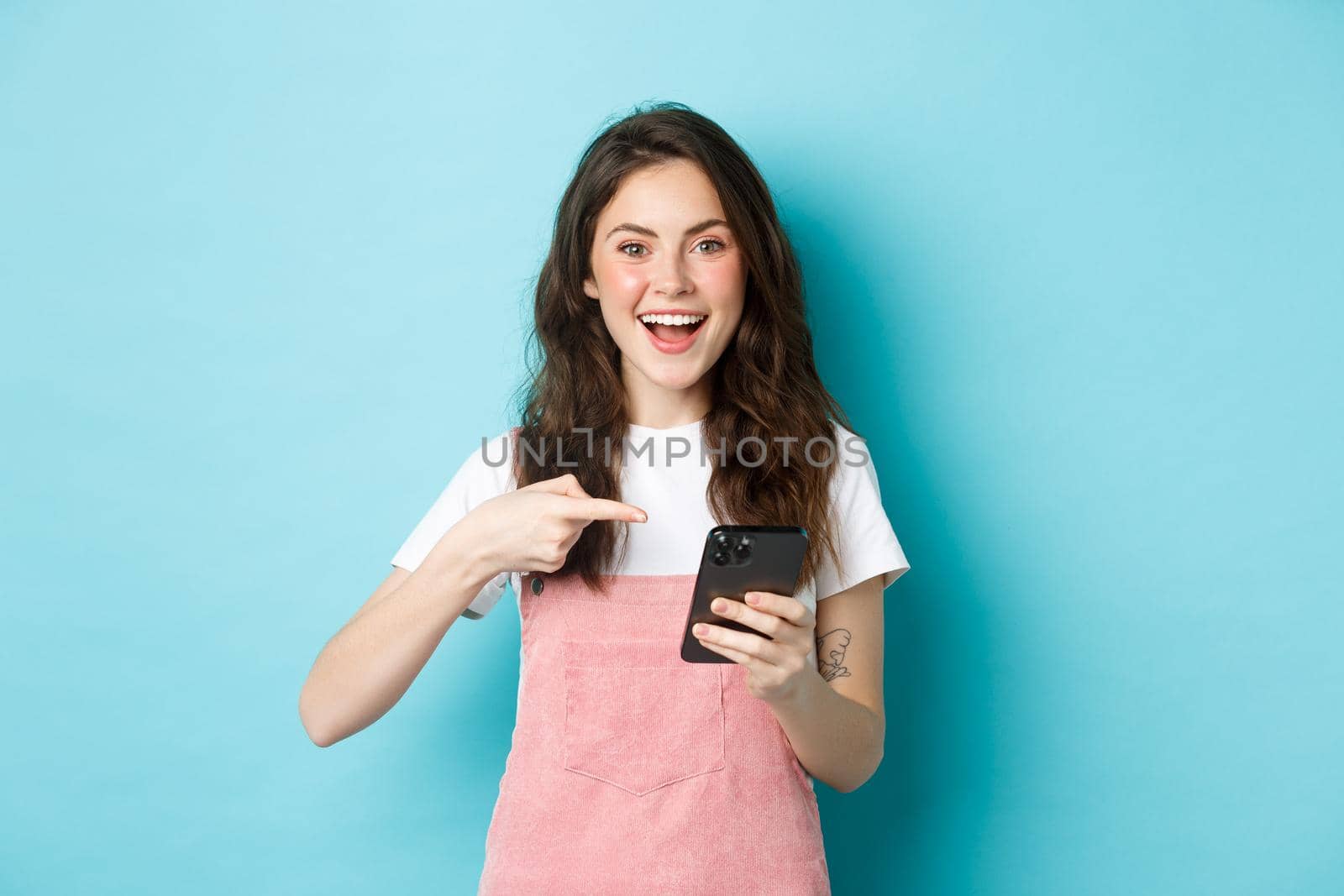 Portrait of cute and cheerful girl pointing finger at her phone, using smartphone shopping app, standing over blue background.