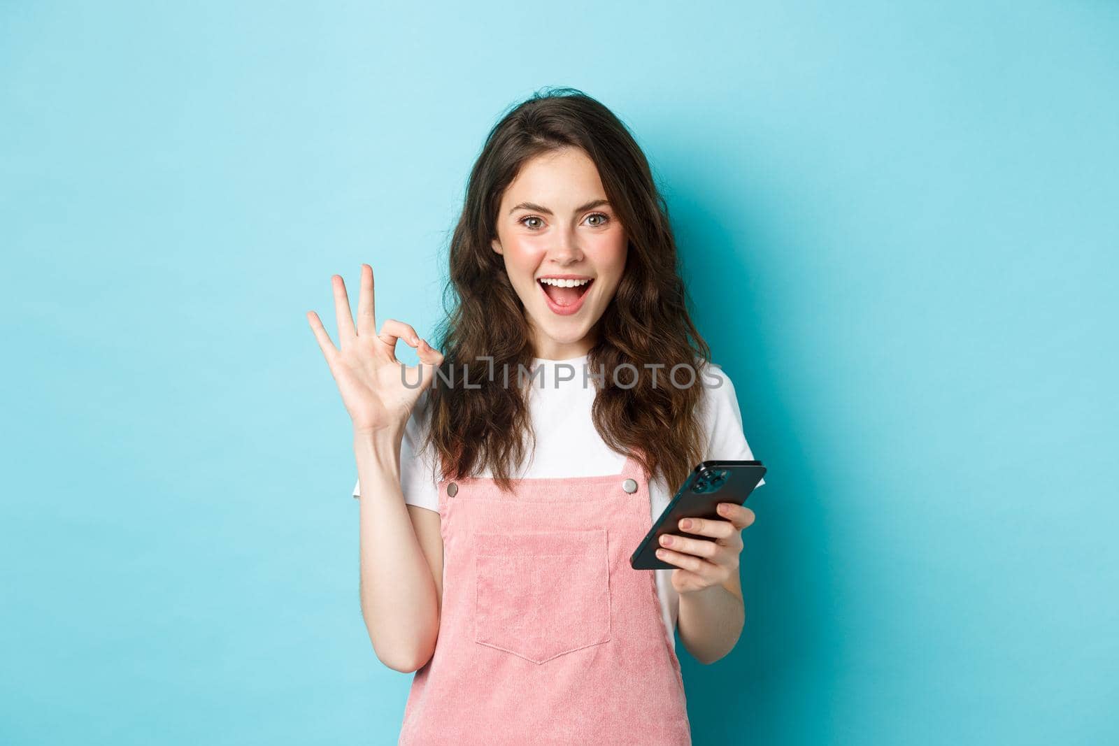 Image of excited and pleased young woman say yes after using smartphone app, online shopping with mobile phone and showing OK sign, standing over blue background by Benzoix