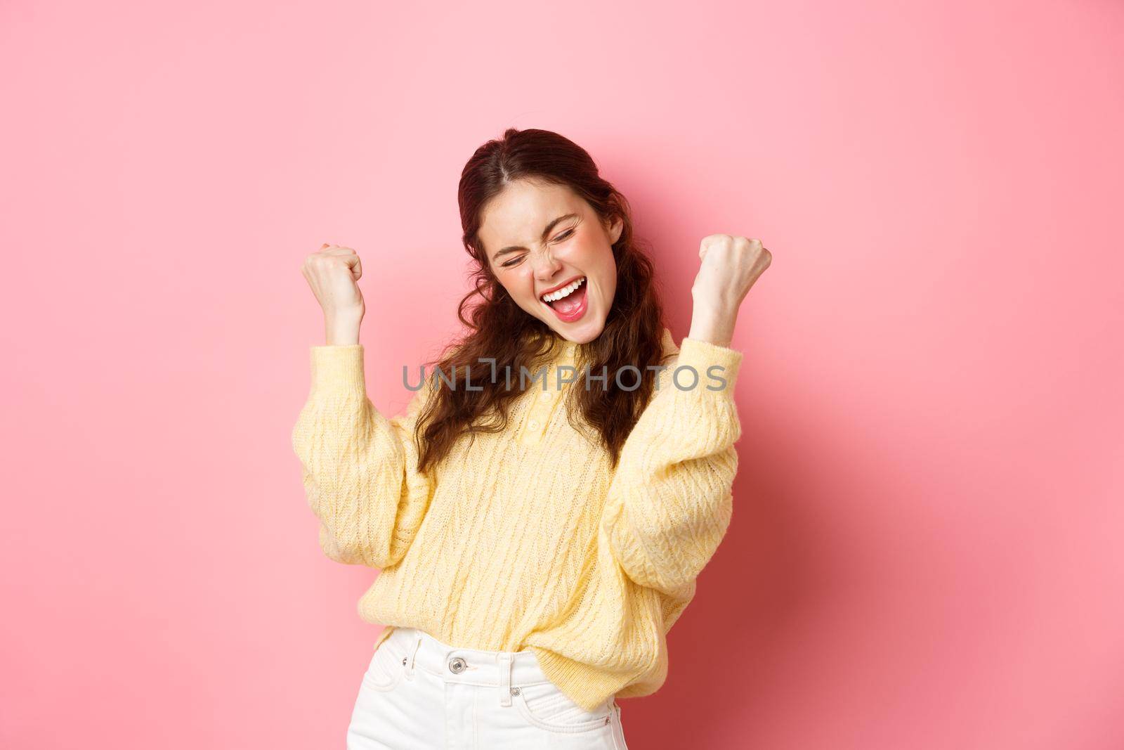 Girl screams with joy and fist pump, say yes, achieve goal or success, celebrating achievement, triumphing and winning, standing over pink background.