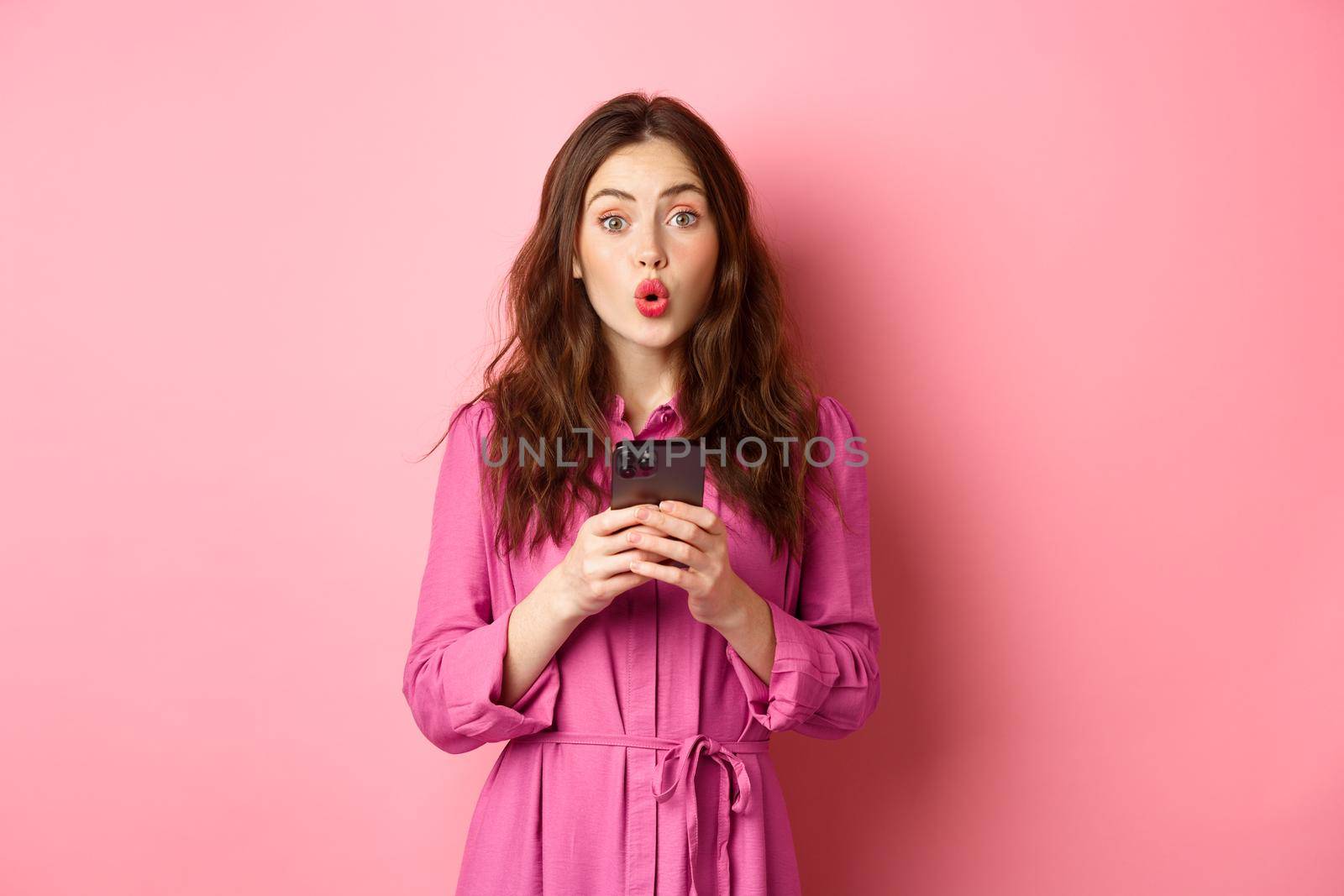 Image of surprised young woman gasping and saying wow, staring impressed at camera after reading news online on mobile phone, standing with smartphone against pink background. Copy space