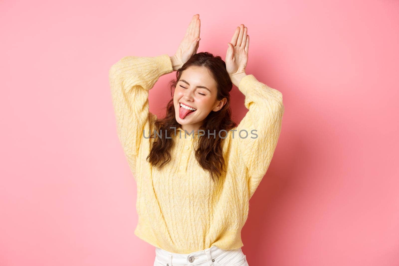 Portrait of funny and cute young woman showing tongue, smiling happy, making easter bunny gesture, standing against pink background.