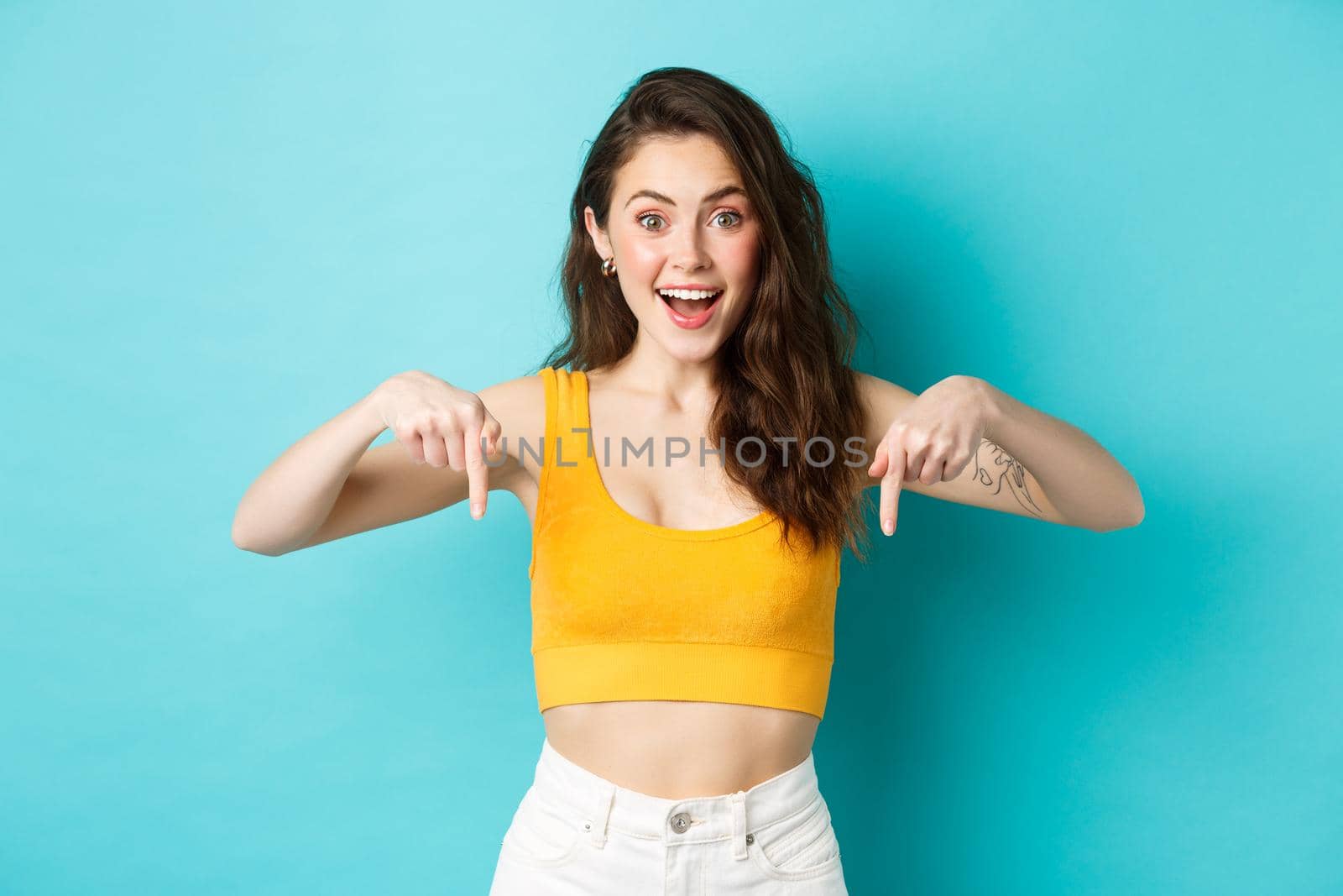 Young happy woman showing awesome news, pointing fingers down, demonstrate advertisement on bottom smiling excited, standing against blue background.