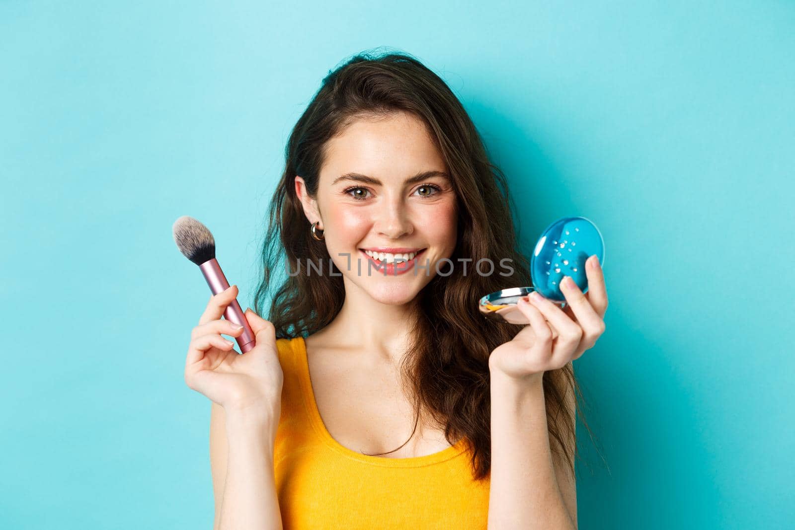 Beauty. Close up of attractive stylish woman applying make up, holding brush and cute pocket mirror, smiling satisfied at camera, standing over blue background.