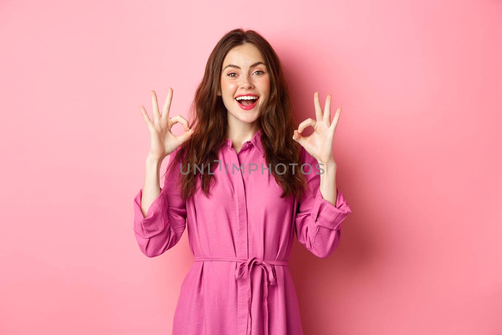 Beautiful woman 20s years, showing okay signs in approval, nod and smile pleased, standing satisfied, standing over pink background.