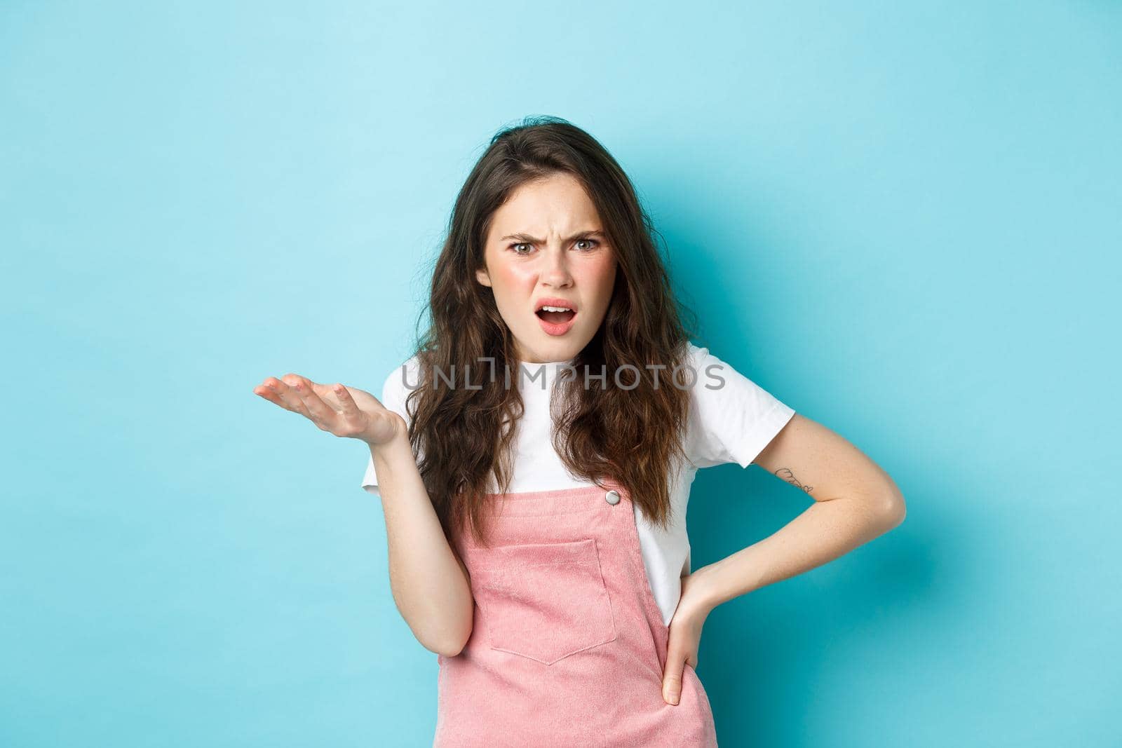 Image of confused and annoyed young woman raising hand and pointing at something terrible, staring puzzled at camera with pissed-off face, standing over blue background.