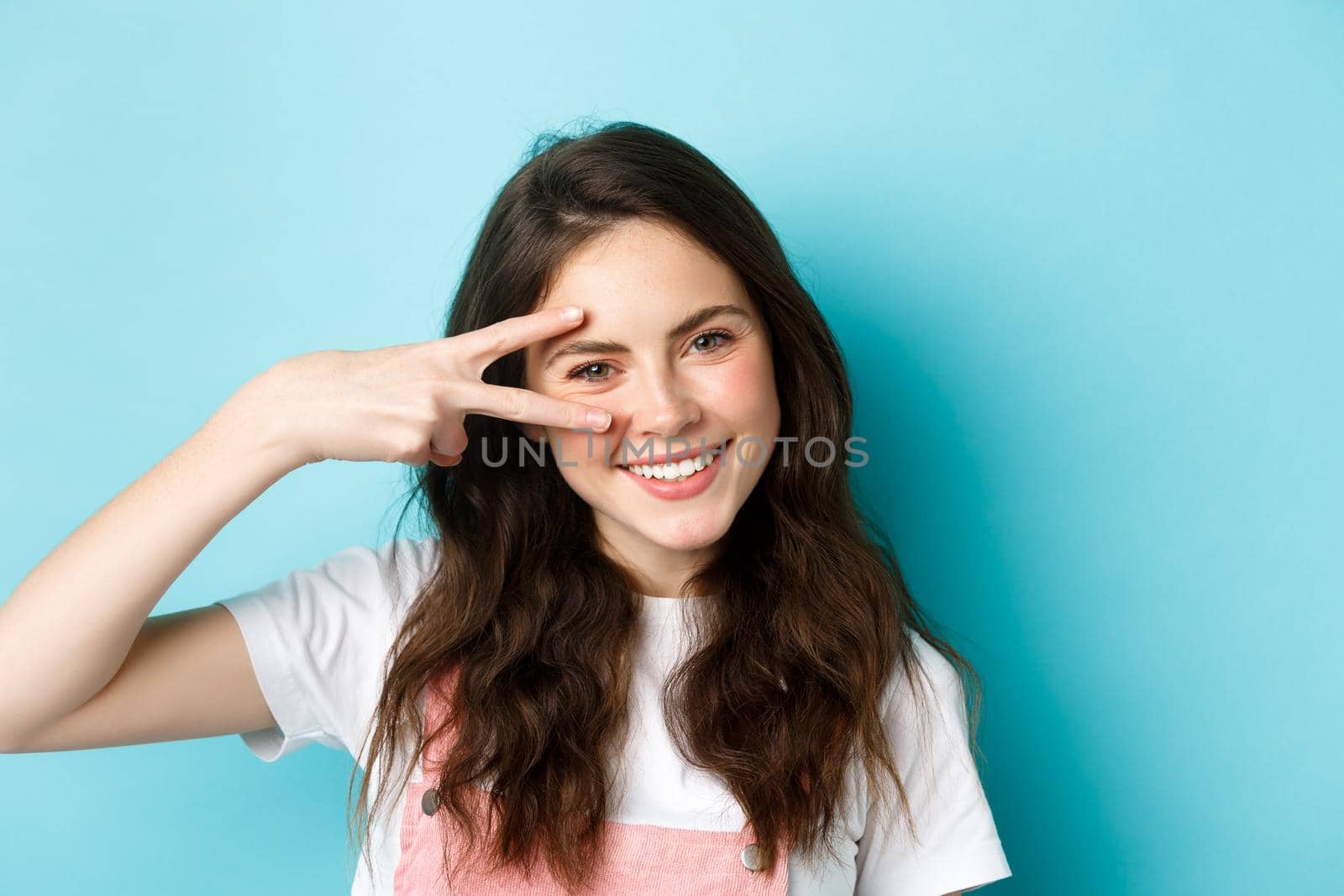 Beauty. Close up of attractive young woman with clean skin and cute blush, showing v-sign near face, smiling with teeth, standing over blue background.