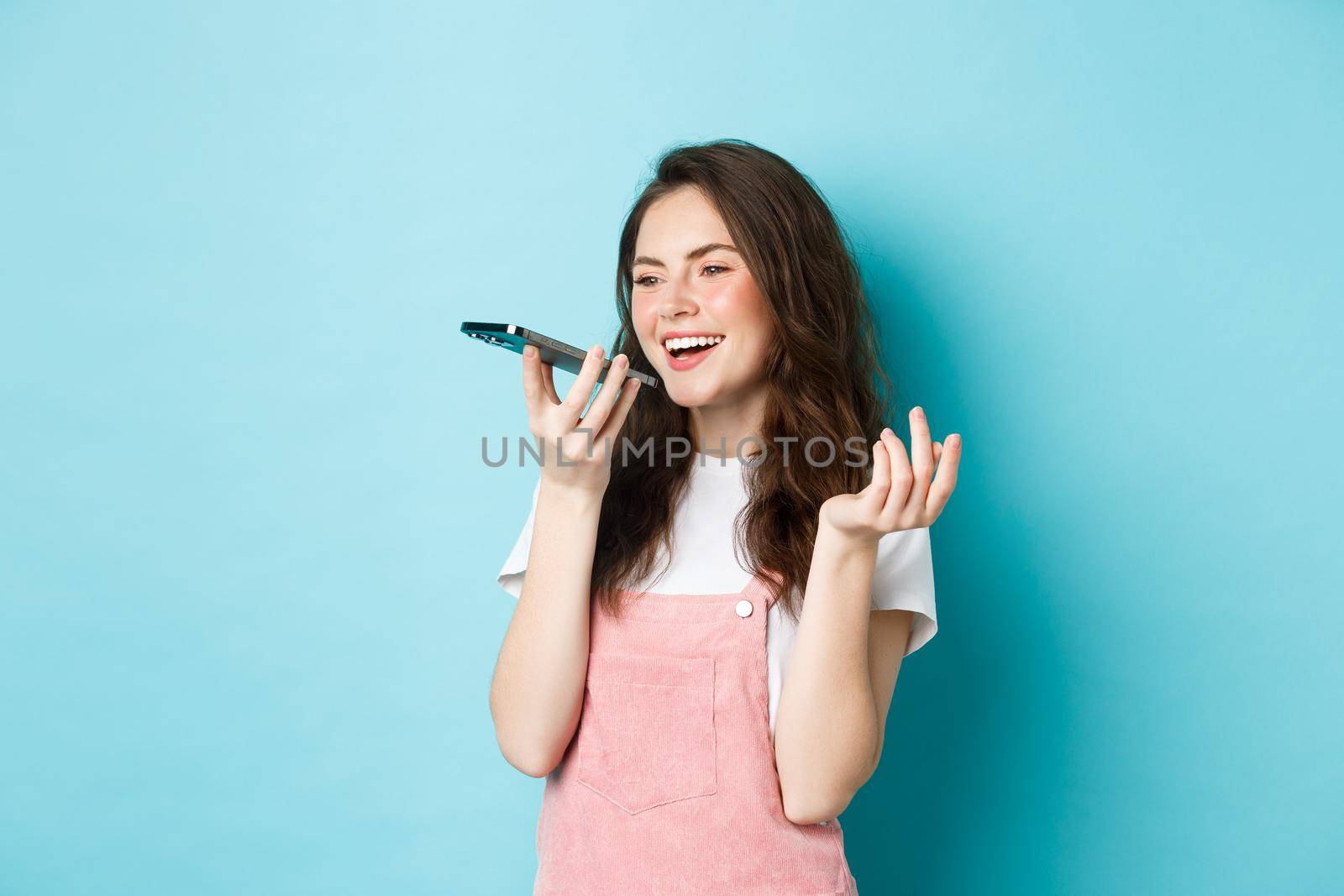 Portrait of smiling woman holding phone near lips and talking, using app translator on smartphone or recording voice message, speaking with speakerphone, standing over blue background by Benzoix