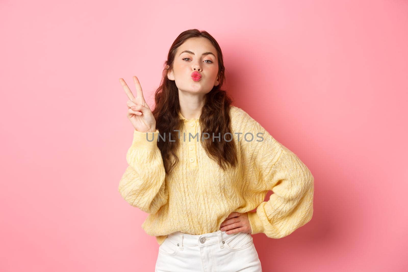 Young glamour woman pucker her lips, making kissing face and v-sign, show peace gesture, standing against pink background.