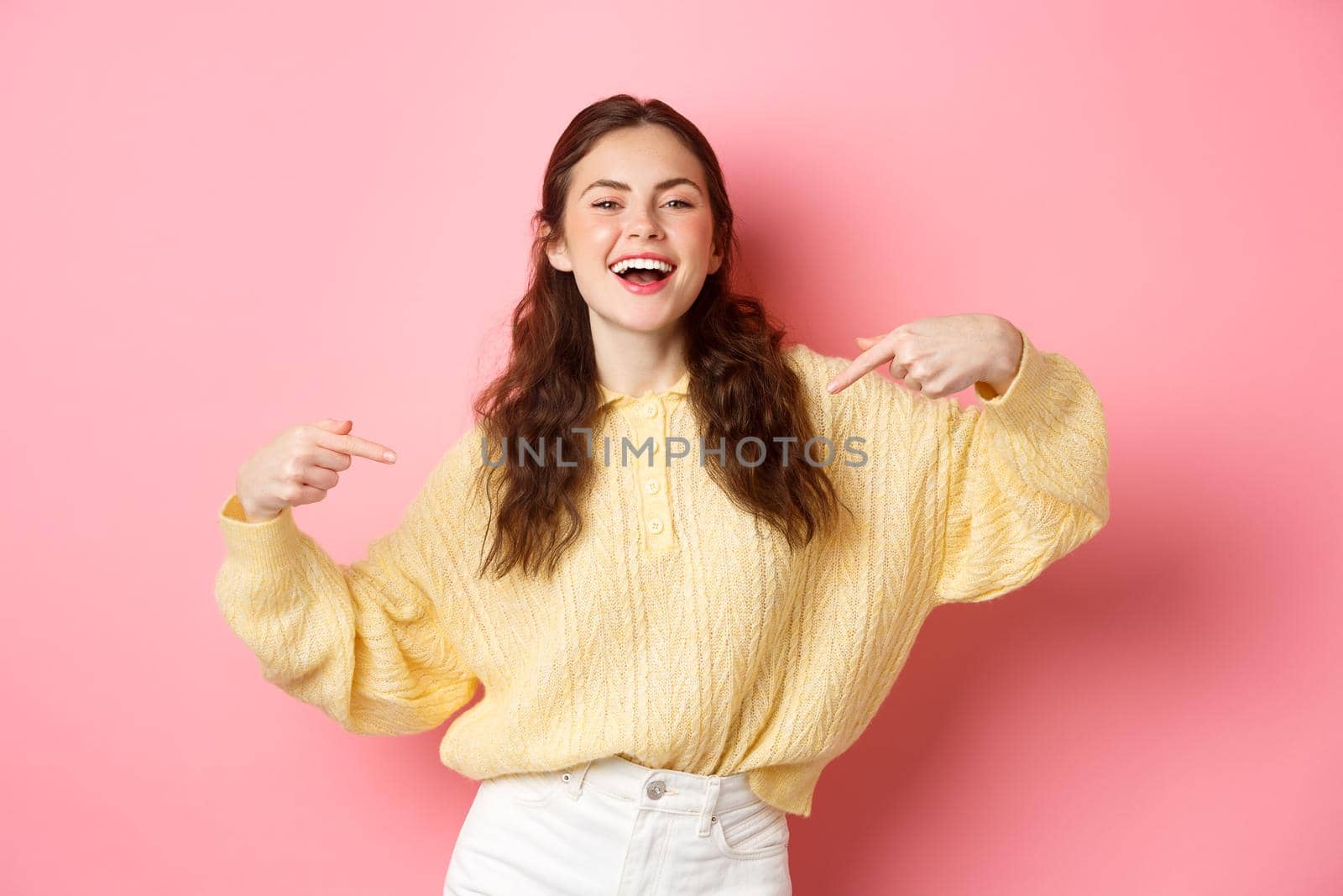 Self-assured happy girl points at herself and laughs, self-promoting and smiling, standing against pink background.
