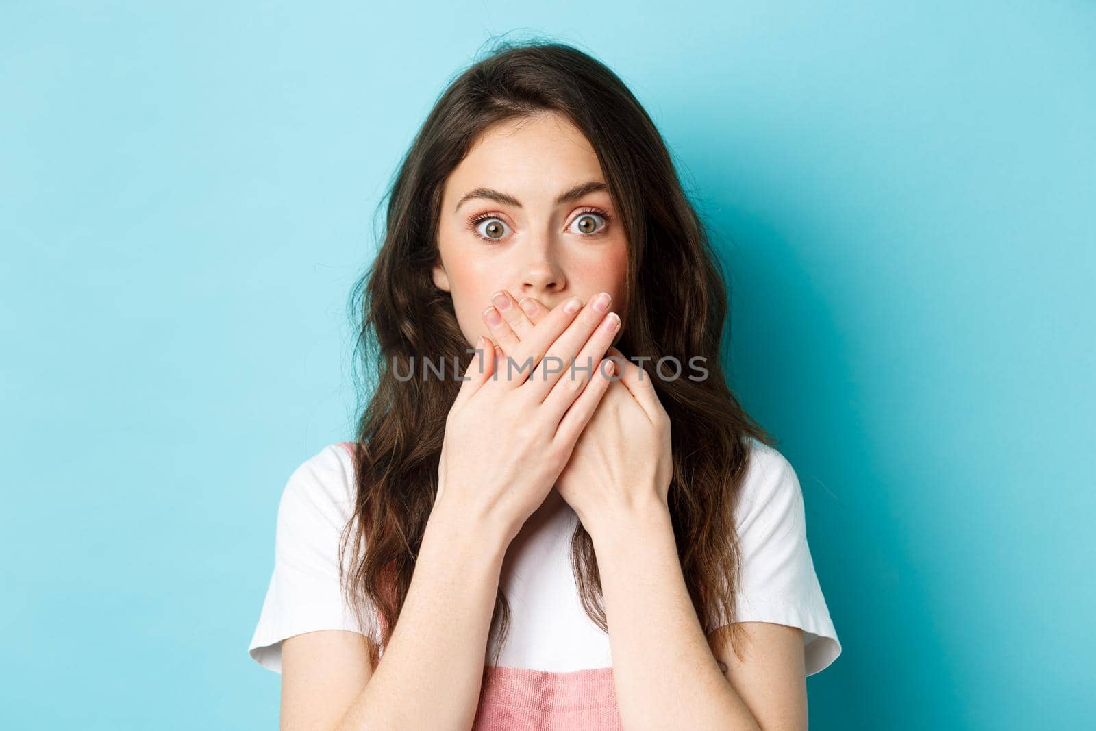 Close up portrait of shocked brunette woman covering hands and stare startled at camera, looking with disbelief and shock, standing against blue background.